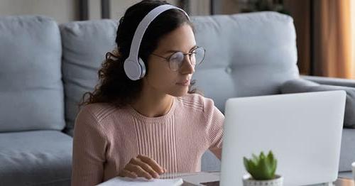 Image of a young female 推荐正规买球平台 student in a pink sweater wearing white headphones looking at her laptop. 